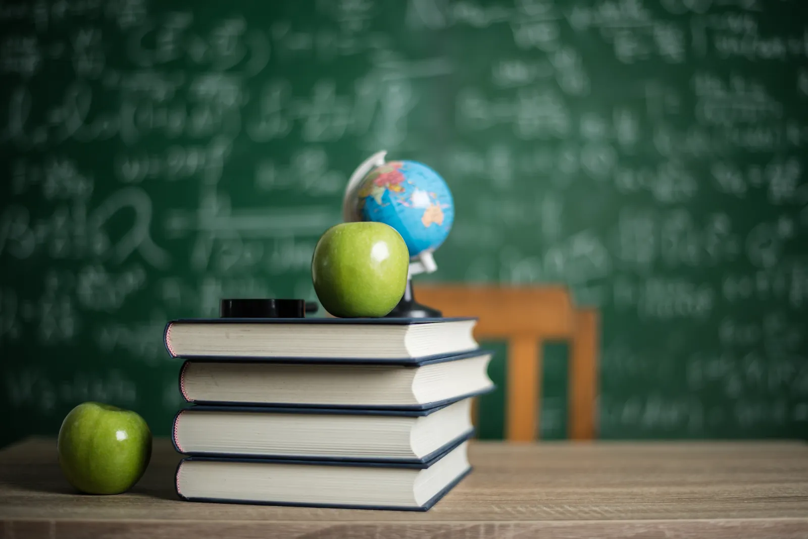 Stack of books, green apples, and a globe in front of a classroom board